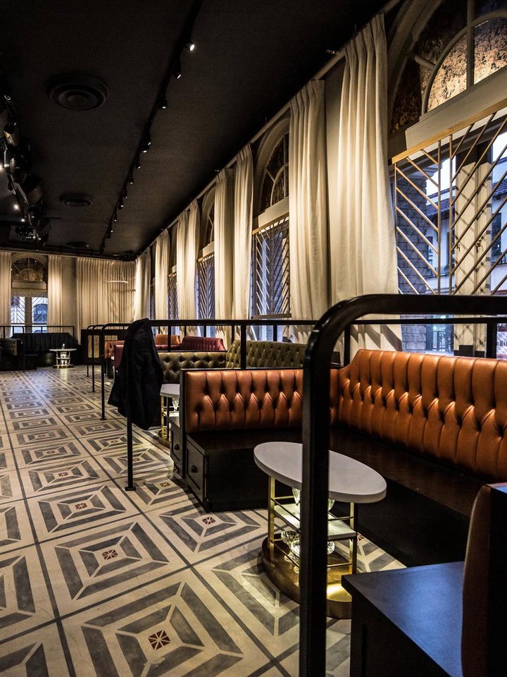 Saint Yves' main lounge, with cognac tufted banquettes and cocktail tables along tall curtained windows and a patterned tile floor